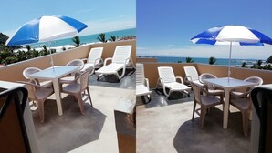 Terraço com cadeiras de estar brancas e uma mesa, um guarda-sol azul e branco, e vista para o mar.