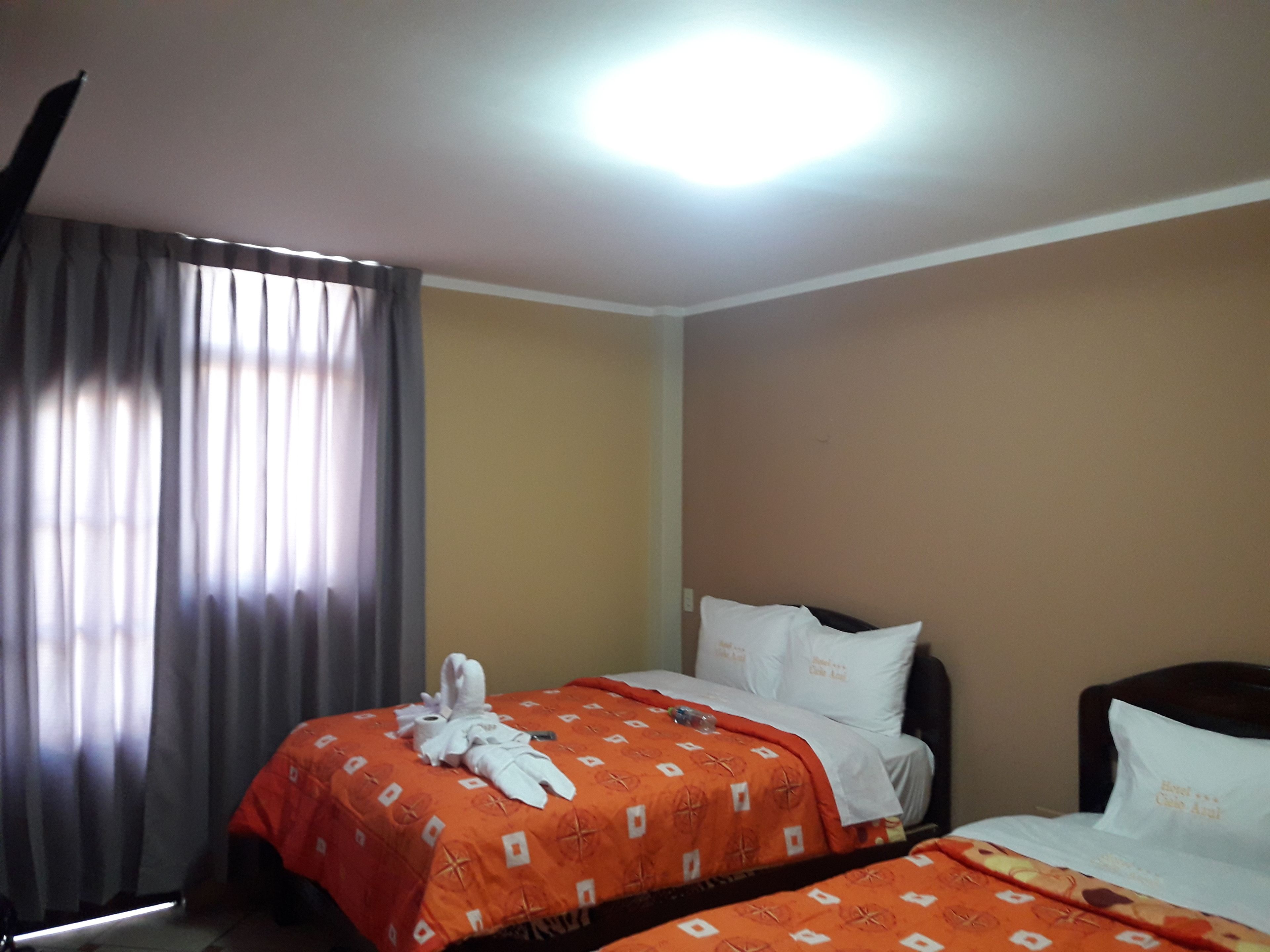 A hotel room with two beds, a large window with curtains, and a bright ceiling light.