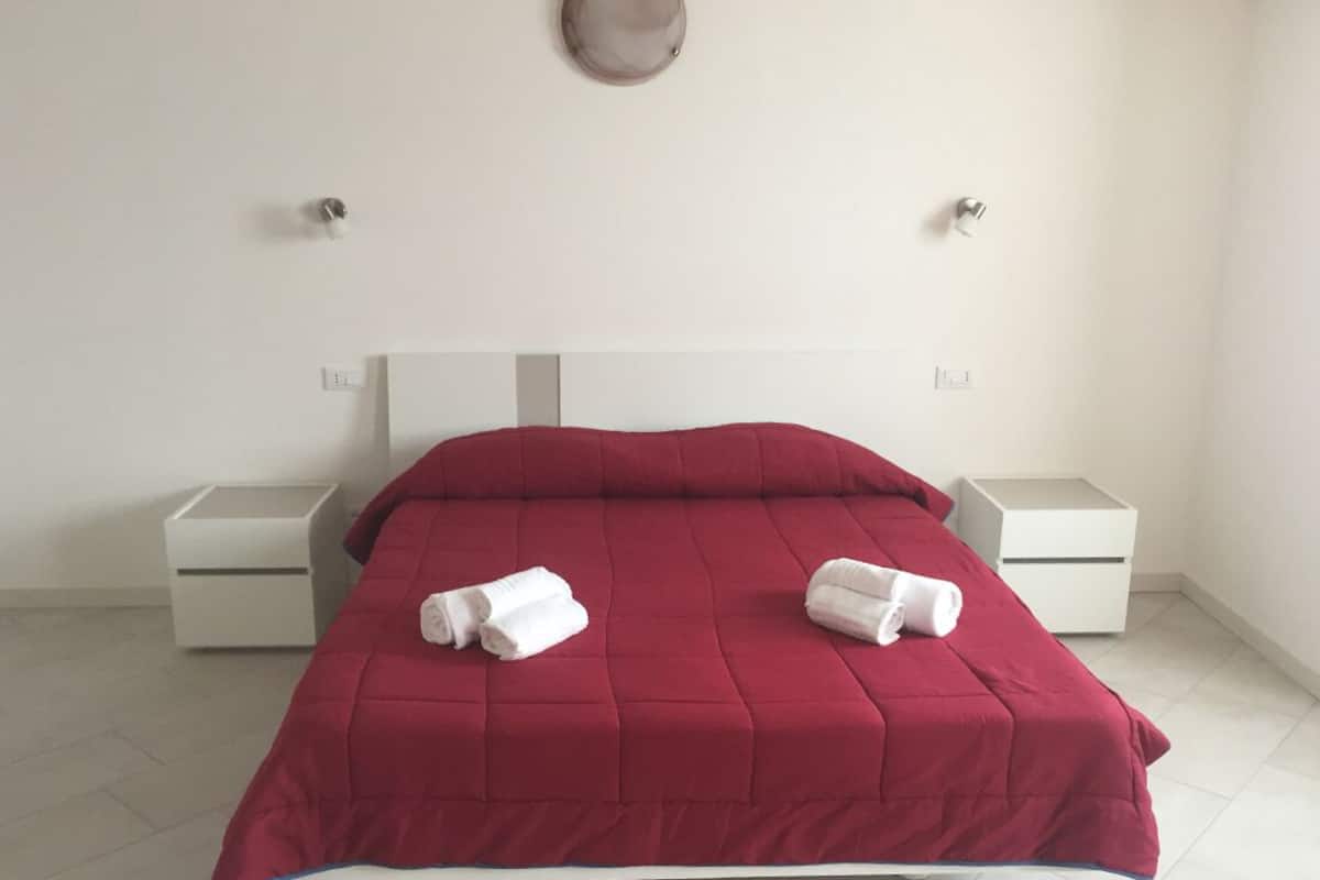A neatly made bed with a red bedspread and two white pillows, accompanied by two white bedside tables.