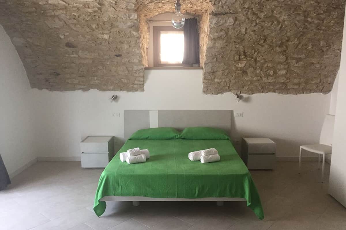 A bedroom with a stone ceiling, a single window, a green bedspread, two bedside tables, and two rolled towels.