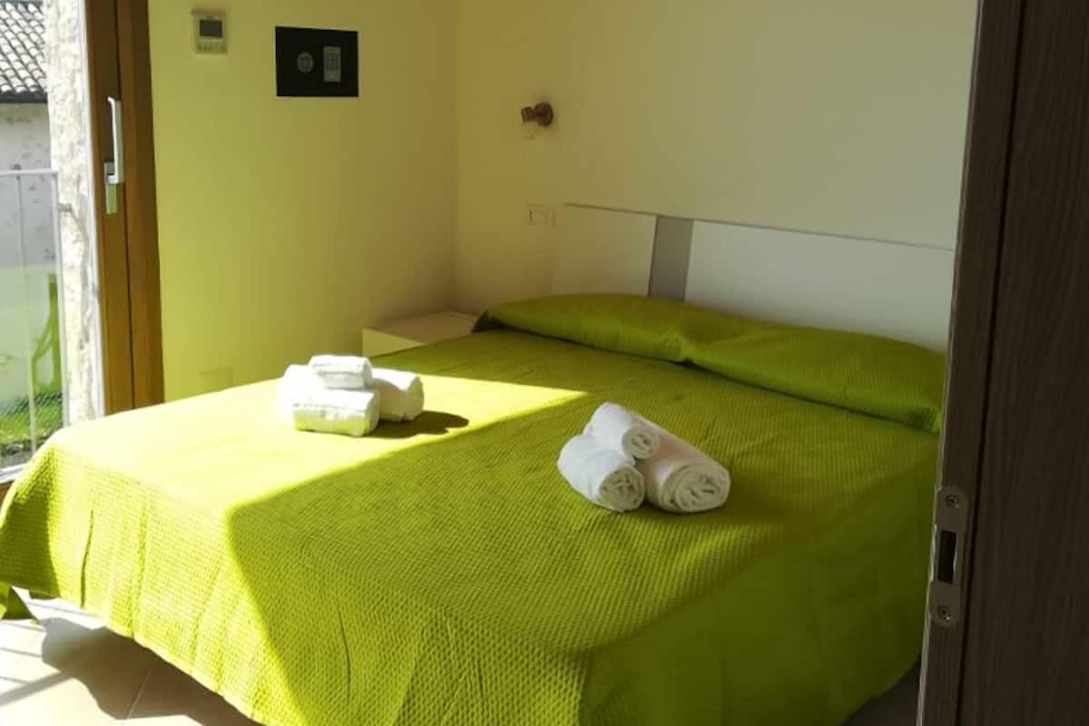 A room with a bed, a door, a light switch, and a small wall-mounted device.