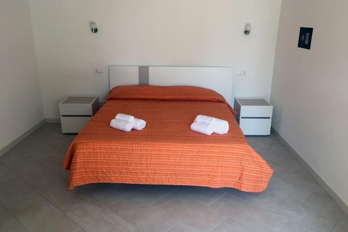 A neatly made bed with an orange bedspread and two white towels, two bedside tables, and a light switch on the wall.