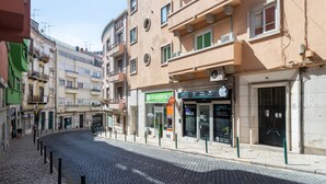 Uma rua de paralelepípedos com edifícios em ambos os lados, incluindo um caixa eletrônico visível e uma loja com uma placa que diz 'Lavandaria'.