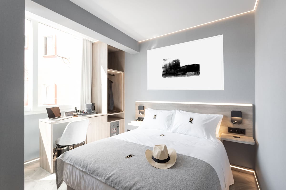 A modern hotel room with a bed, a desk, a chair, a laptop, and a hat on the bed.