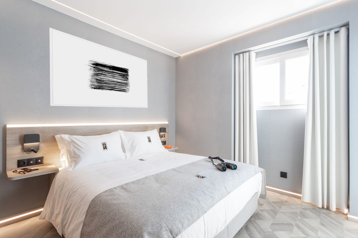A modern hotel room with a large bed, a bedside table with a lamp, a wall-mounted artwork, and a window with curtains.