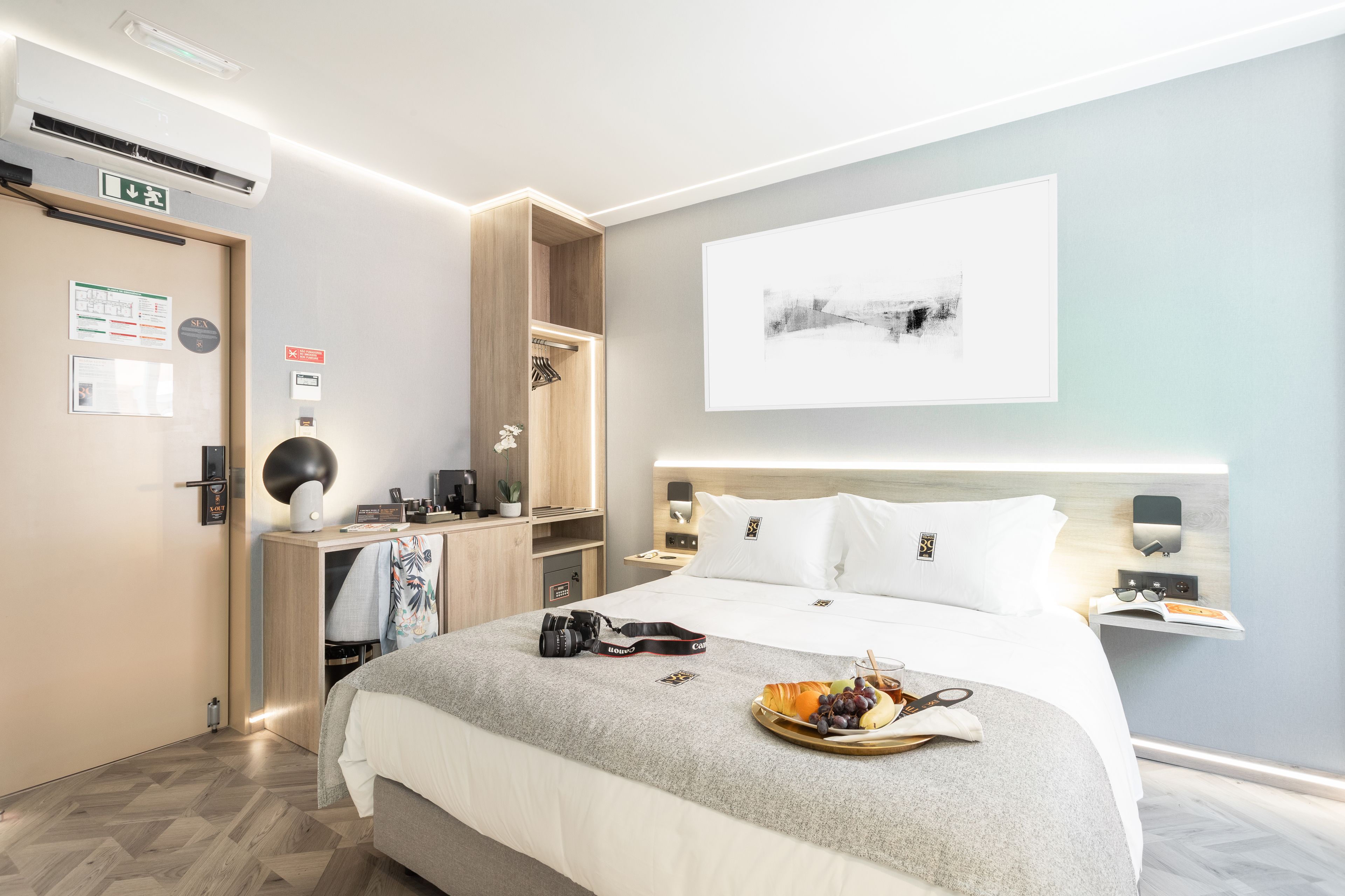 Quarto de hotel moderno com uma cama grande, mesas de cabeceira, uma escrivaninha com luminária, uma obra de arte na parede e uma bandeja com frutas e lanches sobre a cama.