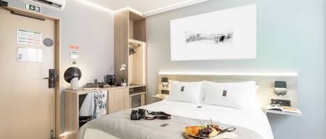 Quarto de hotel moderno com uma cama grande, mesas de cabeceira, uma escrivaninha com luminária, uma obra de arte na parede e uma bandeja com frutas e lanches sobre a cama.