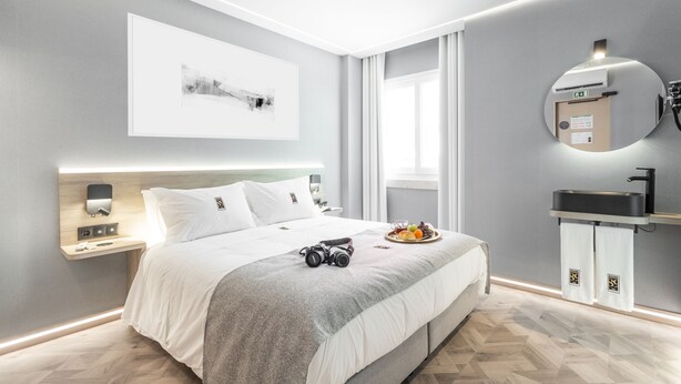 Quarto de hotel moderno com cama grande, mesa de cabeceira com abajur, espelho fixado na parede e janela com cortinas.