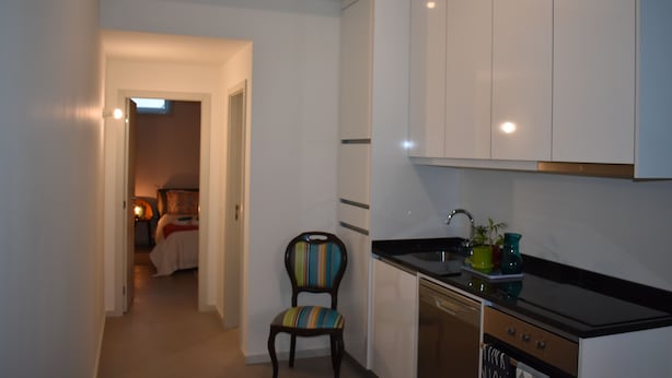 A compact kitchen with white cabinets, a black countertop, and a single chair. A hallway leads to a bedroom with a bed and a nightstand.
