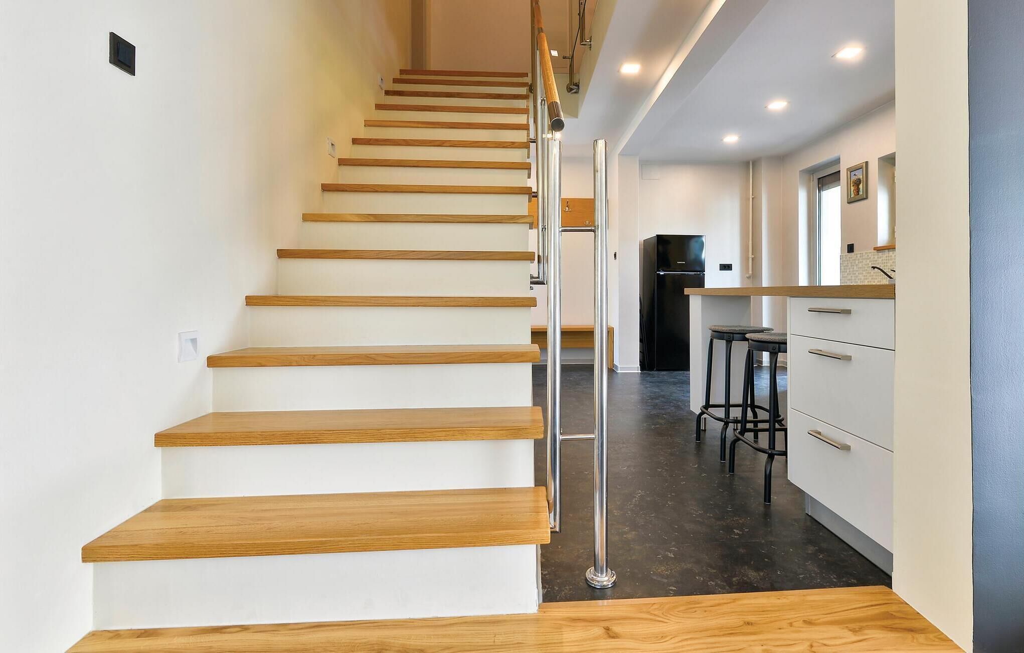 Eine moderne Treppe mit Holzdielen und Metallgeländern führt zu einem Küchenbereich mit weißen Schränken und einem schwarzen Kühlschrank.