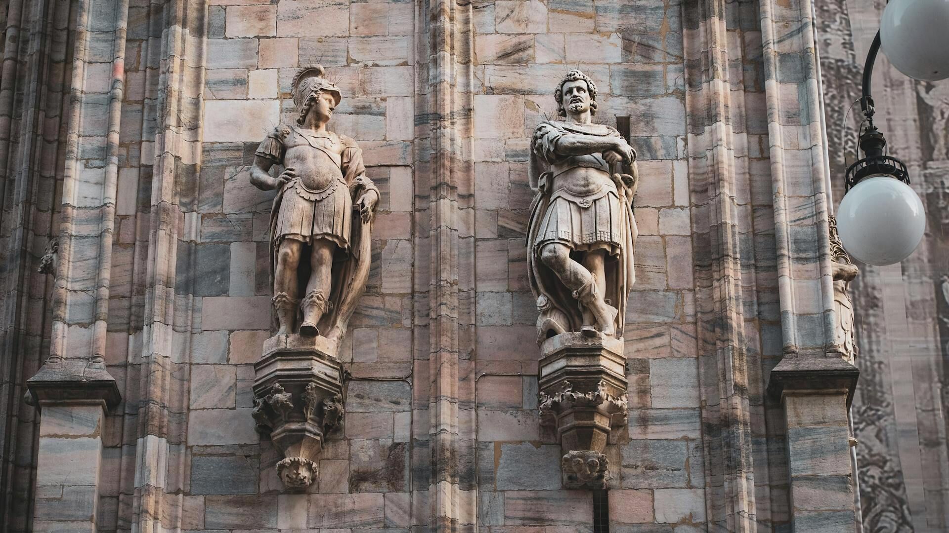 Une façade en pierre ornée de deux statues représentant des personnages vêtus à la mode classique, flanquée de colonnes décoratives et d’un lampadaire.