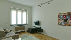 A modern living room with a sofa, a glass dining table, a flat-screen TV, and a large window with blinds. - Apartment Near Corso Buenos Aires, Milano Central (Milan)