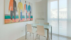 Een moderne eetruimte met een glazen tafel en stoelen, een groot abstract schilderij aan de muur en een groot raam met uitzicht op gebouwen.