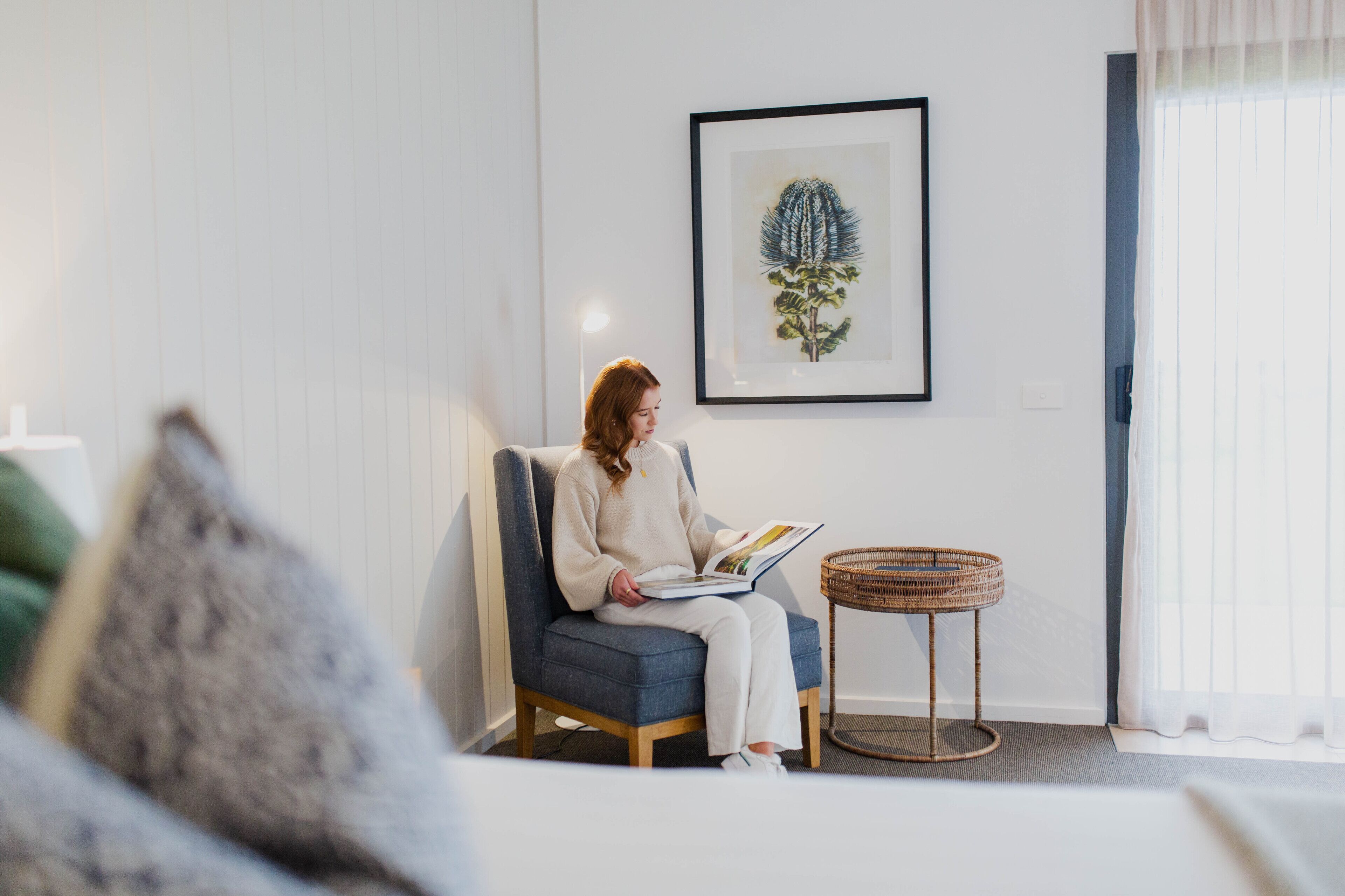 A person sitting on a blue chair reading a book in a room with a framed plant on the wall, a wicker side table, and a window with curtains.