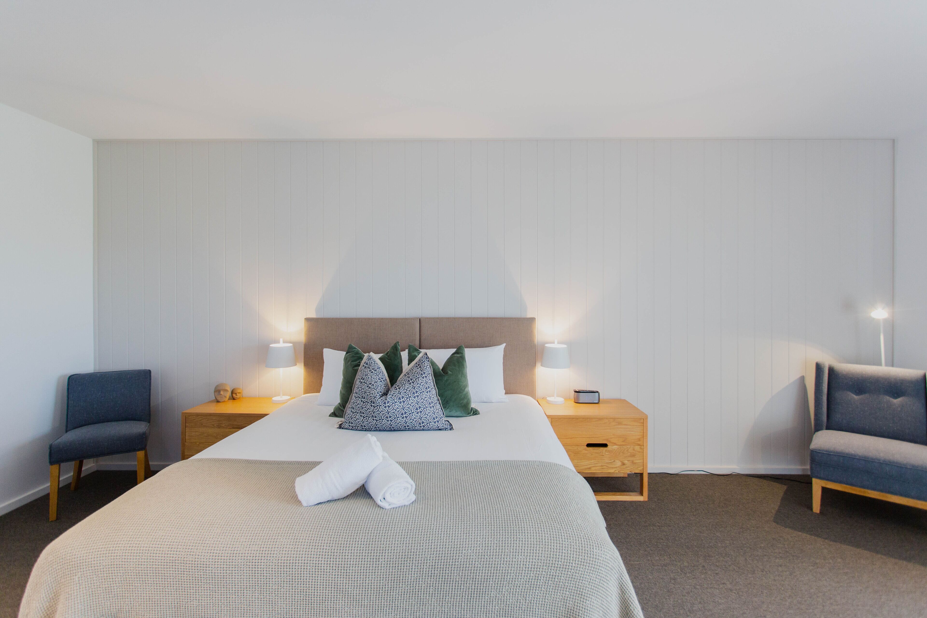 A hotel room with a large bed, two bedside tables with lamps, a chair, and a wooden headboard.