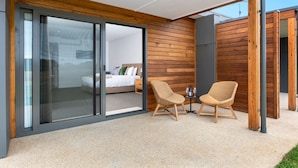 Una habitación moderna con un gran ventanal corredero que da a un dormitorio, dos sillas de mimbre y una mesita con una botella y un vaso.