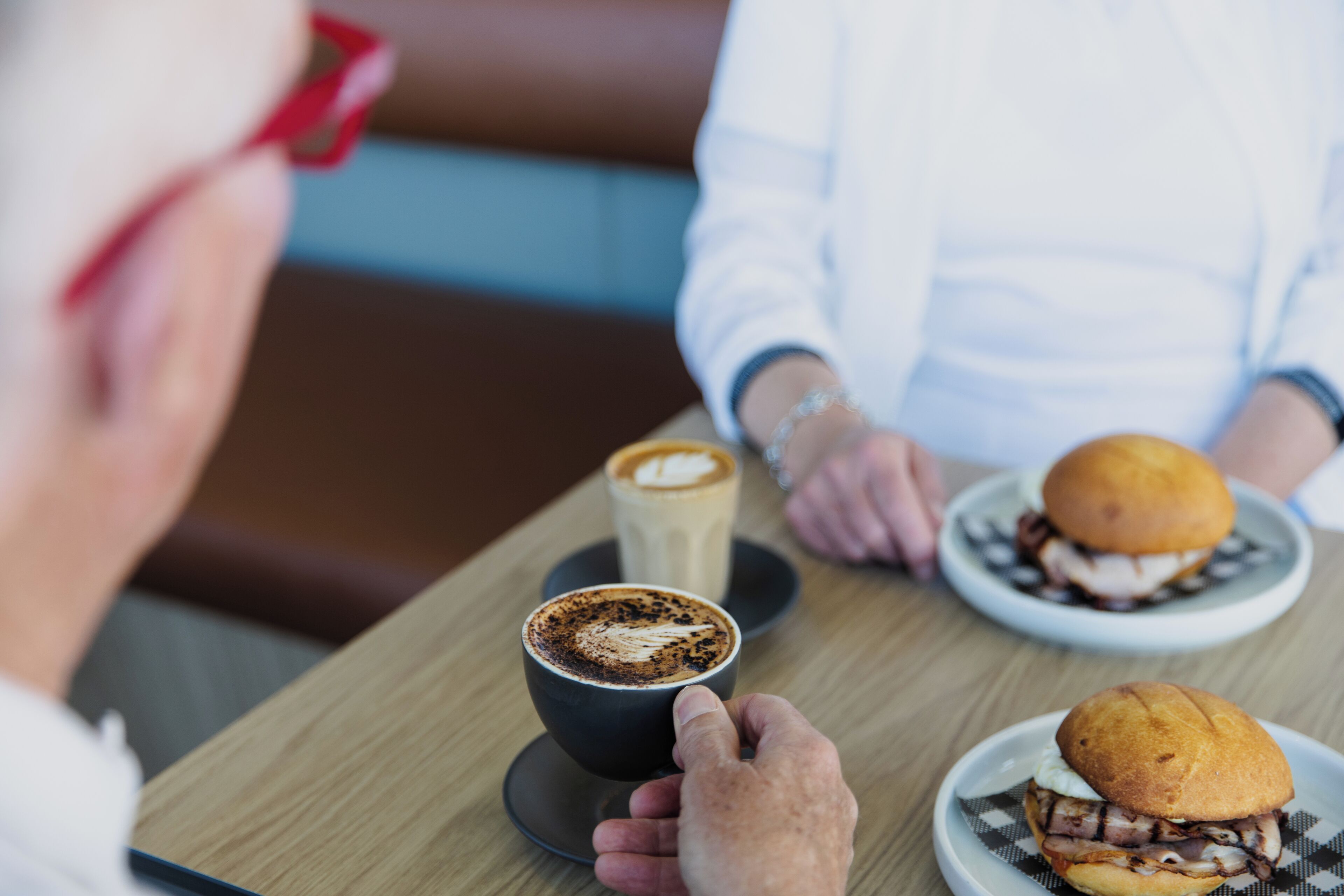 A person holding a cup of coffee with a latte art design, a sandwich on a plate, and another cup of coffee in the background.