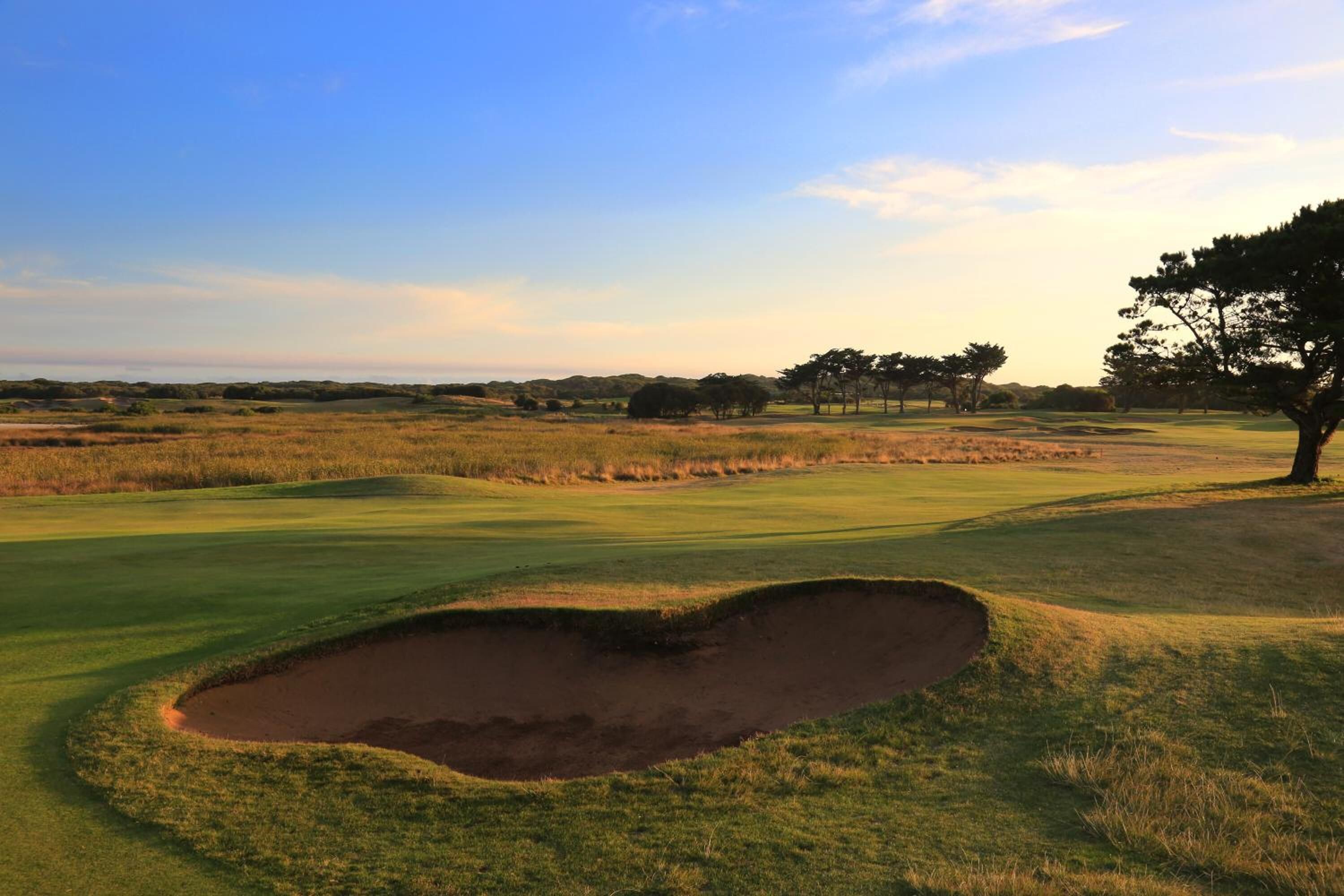 A golf course with a sand bunker, trees, and a clear sky.