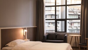 Hotellihuone, jossa on suuri ikkuna, sänky, yöpöytä, tuoli ja pieni pöytä.