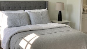 Una cama bien hecha con cabecera gris y ropa de cama blanca, una lámpara de noche y una mesita pequeña.