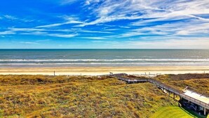 Un paisaje costero con playa, dunas cubiertas de hierba y un paseo marítimo de madera que lleva a una construcción con vistas al océano.