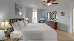 Habitación de hotel con una cama grande, ventilador de techo, televisor y balcón con vista al océano.