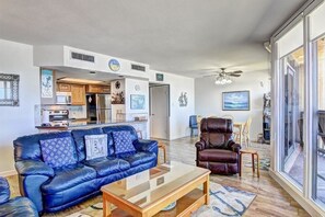Un salon avec un canapé en cuir bleu, une table basse en bois et un fauteuil en cuir marron. Au fond, on aperçoit une cuisine équipée d’un four à micro-ondes, d’un four et d’un réfrigérateur.