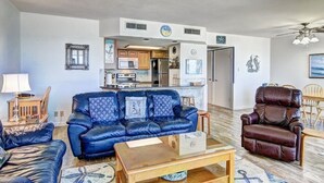 Een woonkamer met een blauwe leren bank, een bruine lederen fauteuil, een houten salontafel en een keuken in de achtergrond.