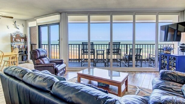 Een ruime woonkamer met een grote schuifpui die uitkomt op een balkon met uitzicht op het strand.