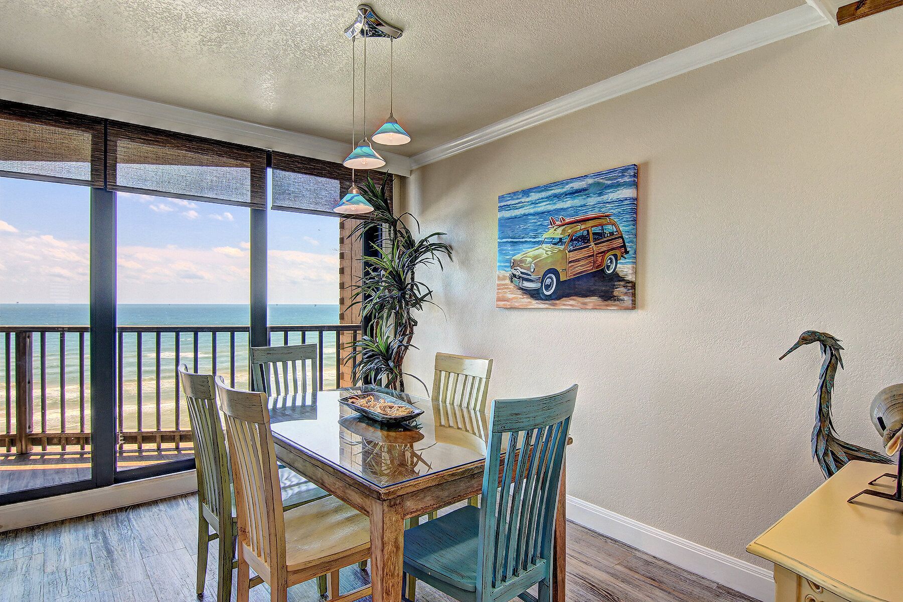 A dining area with a view of the ocean, a painting of a car on the wall, and a bird sculpture.