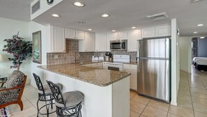 Una cocina moderna con armarios blancos, encimeras de granito, electrodomésticos de acero inoxidable y una isla central con taburetes de bar.