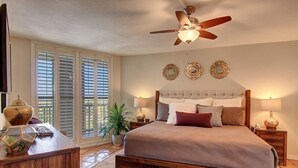 Une chambre à coucher avec un grand lit, une tête de lit en bois, deux tables de chevet avec des lampes, un ventilateur de plafond et une fenêtre avec des stores.