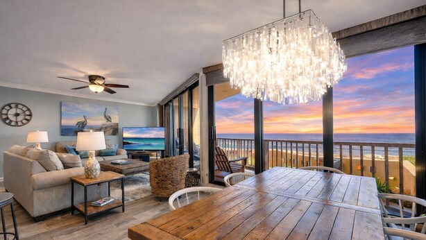 Amplia sala de estar con una mesa de comedor de madera, asientos cómodos, un televisor grande y un balcón con vista al atardecer sobre el océano.