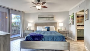 Un dormitorio moderno con una cama grande, dos mesitas de noche con lámparas, un ventilador de techo, vistas al exterior y un baño visible a través de una puerta abierta.