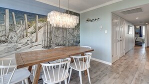 Comedor con una mesa de madera y sillas blancas, un mural decorativo y un pasillo que conduce a los dormitorios.
