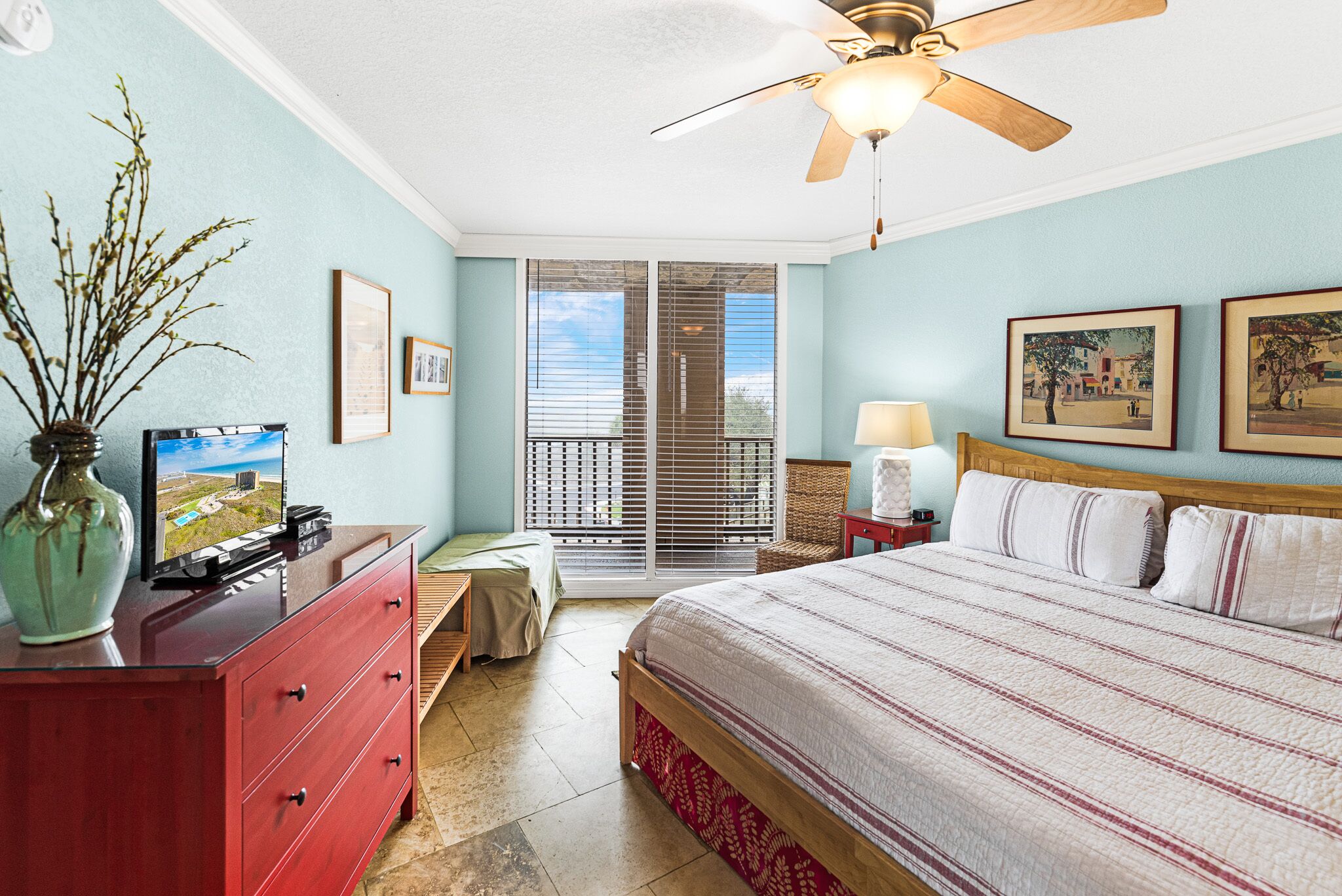 Une chambre à coucher comprenant un lit, une commode, un téléviseur, un ventilateur de plafond et une fenêtre avec des stores.