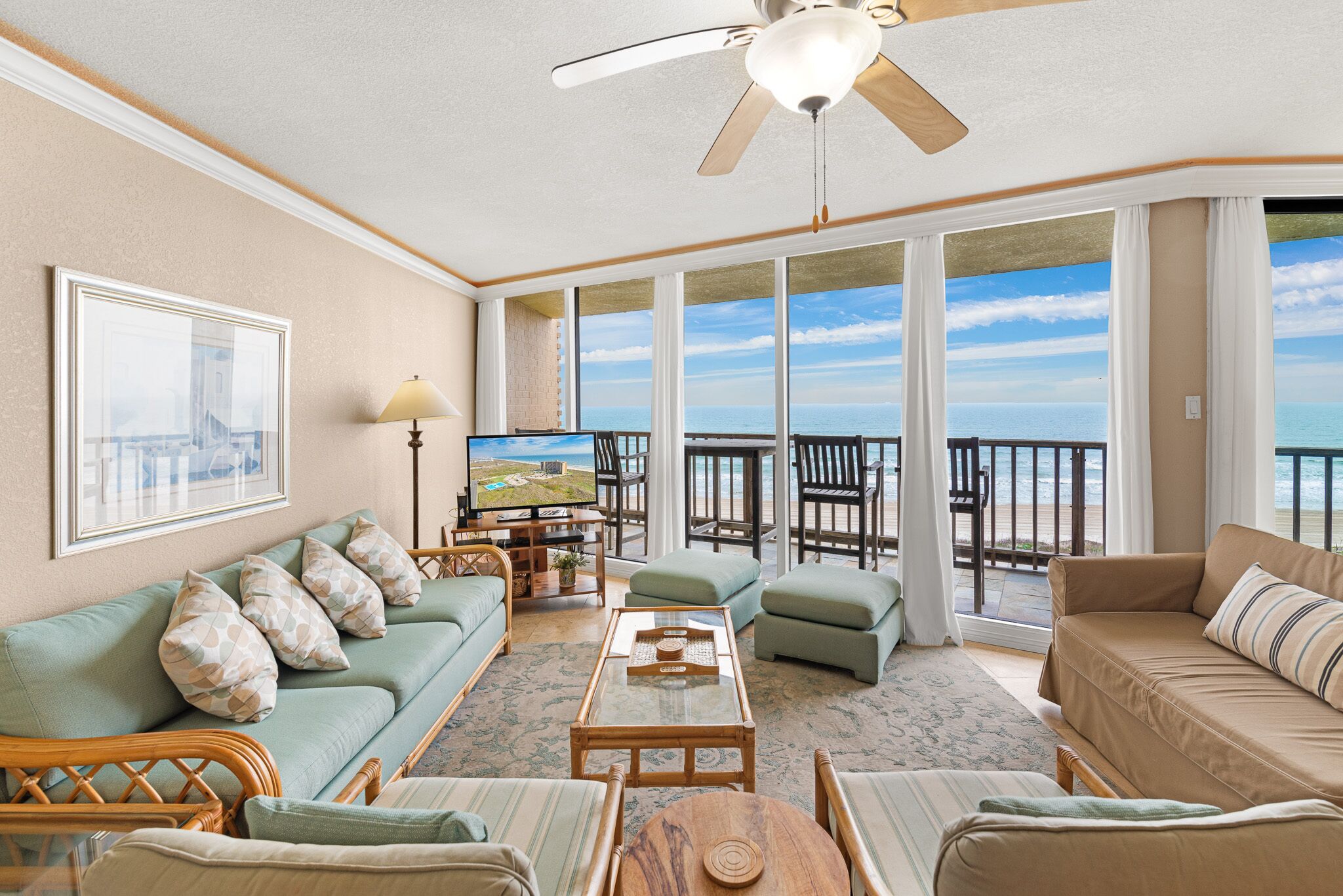 A spacious living room with a large window overlooking the ocean, a ceiling fan, and a television on a wooden coffee table.