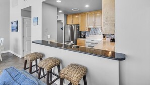 A modern kitchen with wooden cabinets, a stainless steel refrigerator, and a black countertop. There are three woven stools and a blue towel on the floor.