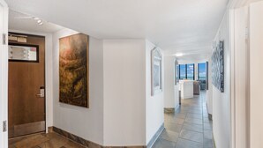 A hallway with a wooden door, a large abstract painting, and a view of a kitchen area with a window. - Sea Gull #1110: Beachfront 3 Bedroom 2.5 Bathroom With Spectacular Views and 24 Hour Management (Port Aransas)
