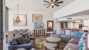 A spacious living room with a large sectional sofa, a coffee table, and a ceiling fan. There are framed artworks on the walls and a dining area in the background. - Sea Gull #1110: Beachfront 3 Bedroom 2.5 Bathroom With Spectacular Views and 24 Hour Management (Port Aransas)