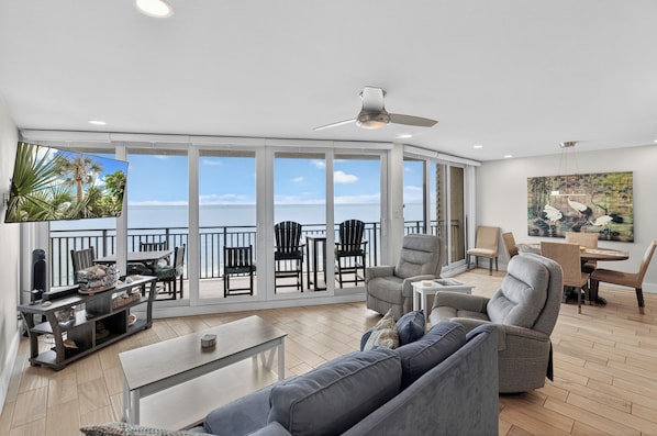 A spacious living area with a balcony, large windows, a ceiling fan, and a flat-screen TV.