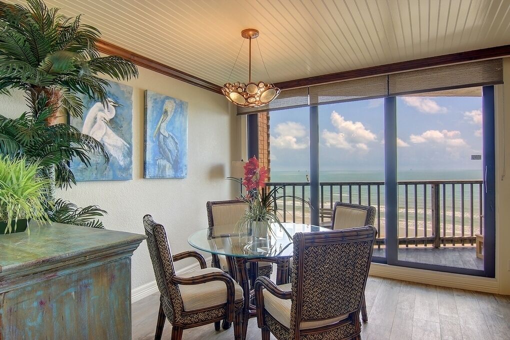 A dining area with a glass table and chairs, a decorative vase, and a view of the beach through large windows.