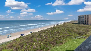Ein Strand mit klarem Himmel, Wellen im Ozean und einer Grünfläche mit einem Gebäude im Hintergrund.