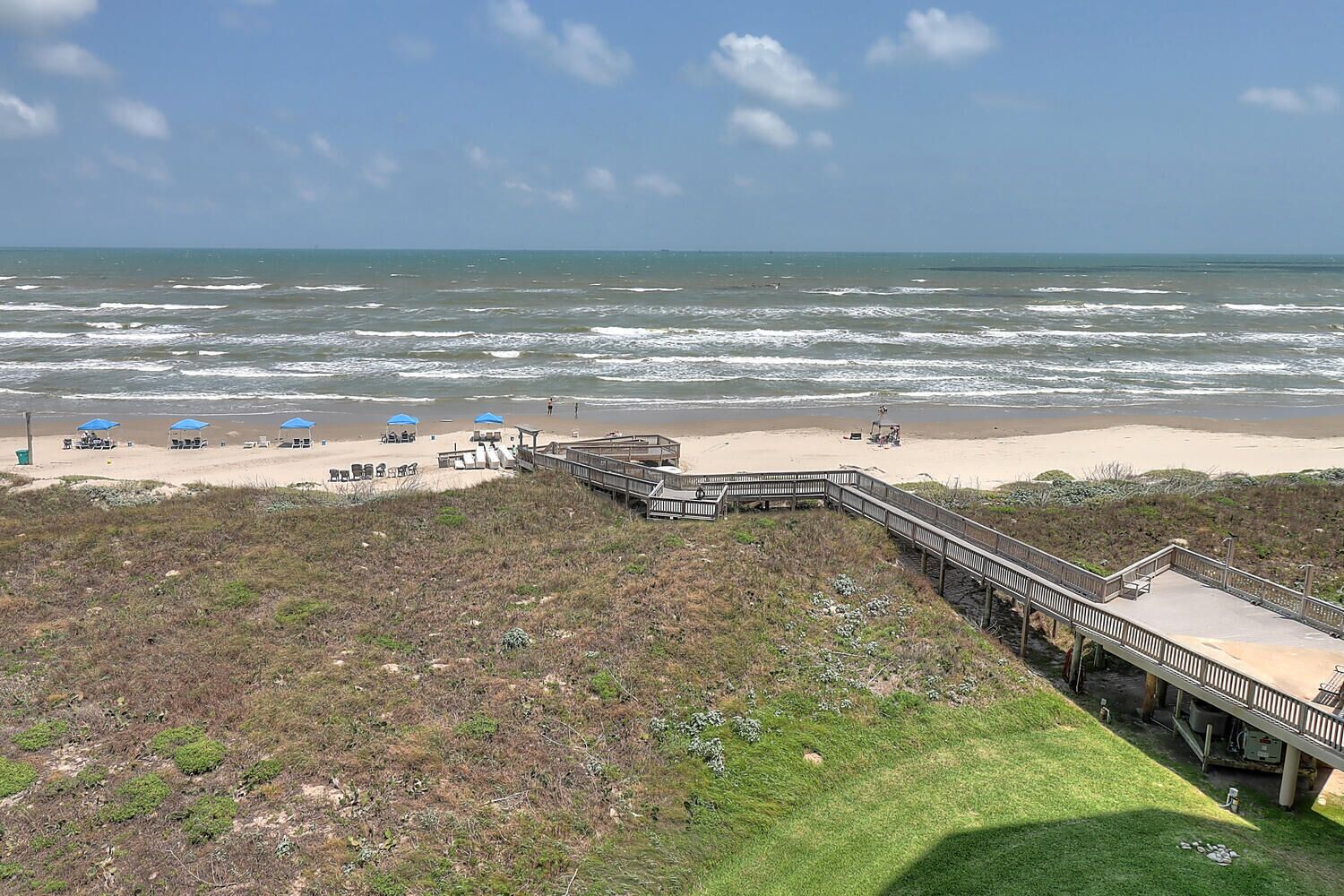 A beach with a wooden boardwalk leading to a sandy area with blue umbrellas and beach chairs.