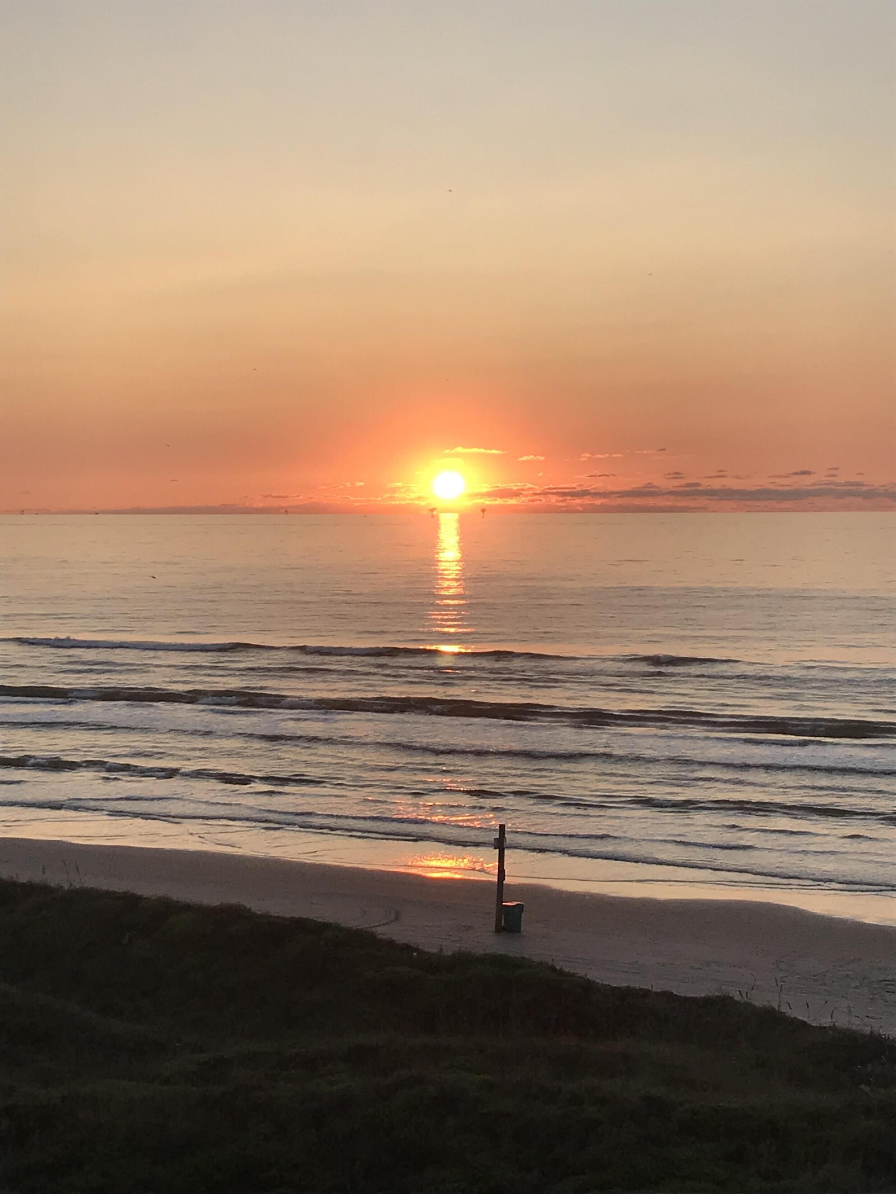 Um pôr do sol sobre um mar calmo, com uma praia de areia e uma pequena estrutura ao primeiro plano.