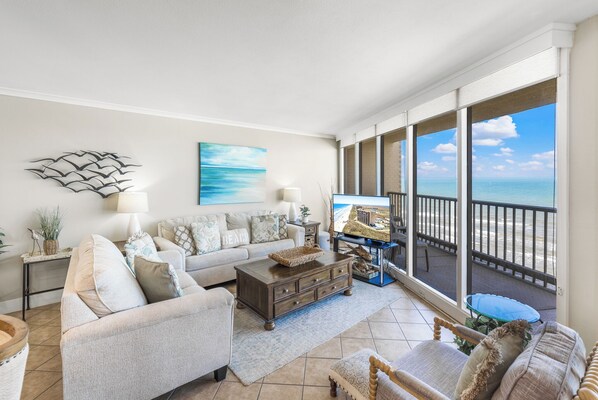 A spacious living room with a large window overlooking the ocean, a comfortable sofa set, a coffee table, and a television.