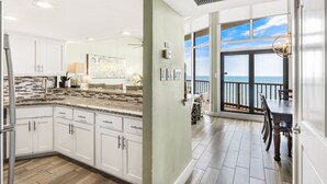 Een moderne keuken met witte kasten, granieten aanrecht en een groot eiland. Er is een eethoek met uitzicht op de oceaan door grote ramen.