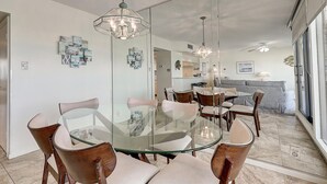 A modern dining area with a glass table and wooden chairs, a chandelier, and a view of the kitchen and living area through a glass partition. - Sea Gull #704: Beachfront 2 Bedroom 2 Bathroom With Spectacular Views and 24 Hour Management (Port Aransas)