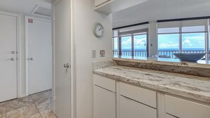 A kitchen with white cabinets, a granite countertop, a clock, and a view of the ocean through large windows. - Sea Gull #704: Beachfront 2 Bedroom 2 Bathroom With Spectacular Views and 24 Hour Management (Port Aransas)
