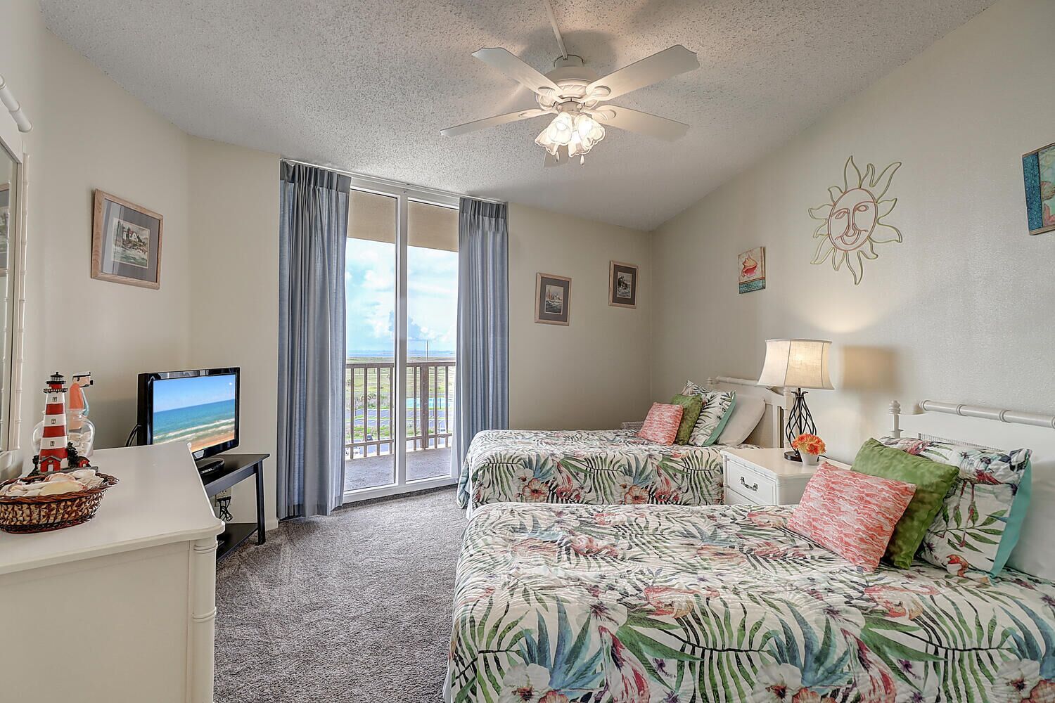 A bedroom with two beds, a desk with a television, a ceiling fan, and a view of the outdoors through a sliding door.